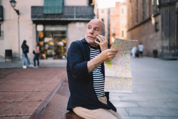 Aged Caucasian tourist reading travel map searching showplace location during international cellphone conversation for discussing vacations trip