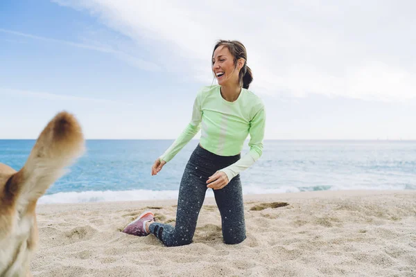 Full body happy slim adult female in activewear and sneakers kneeling on sandy waterfront and laughing while looking at running dog