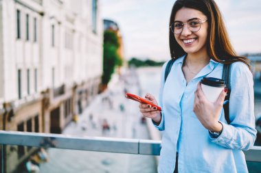 Moda gözlüklü neşeli kadının yarısı kameraya gülümsüyor açık havada cep telefonu kullanıyor.