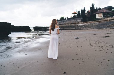 Tanımlanamayan bir kadının arka görüntüsü günlük giysiler içinde kumlu sahilde yürürken dalgalanan deniz kenarında ve tatildeyken başka yere bakarken.