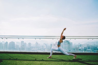 Yan bakışta Virabhadrasana pozu alan genç, ince kadın bulanık arka planda gökdelenin yeşil çatısında yoga yaparken görülüyor.