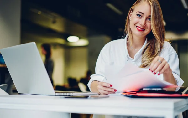 Positive adult lady sitting at table in modern workplace and checking papers while working remotely on laptop and looking at camera