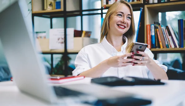 白いシャツを着た陽気な若い女性労働者がノートパソコンを持ってテーブルに座り スマートフォンを手に取りながら見て笑っています — ストック写真