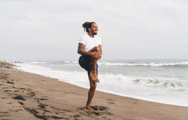 Full body barefoot African American male athlete in sportswear standing on sandy beach and doing stretching exercise during workout in summer