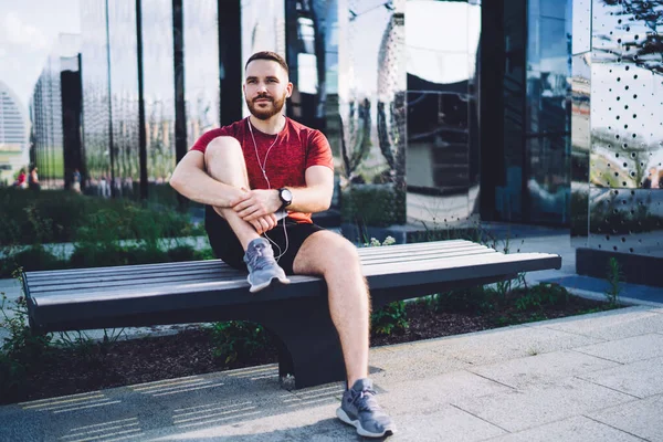 Ciddi sakallı sporcu erkek sporcu. Sokakta bankta otururken müzik dinliyor ve dalgın dalgın bakıyor.