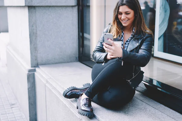 Crop smiling female in casual outfit chilling on concrete windowsill with takeaway coffee while using smartphone communicating with friends on street