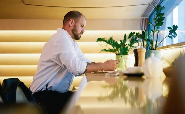 Side view of pensive male in office clothes sitting at table and typing message on smartphone while spending time in cafe