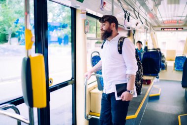 Tablet ve kol saati ile günlük giysiler giymiş genç sakallı bir adam trende dikiliyor ve gündüz gözünü kaçırıyor.