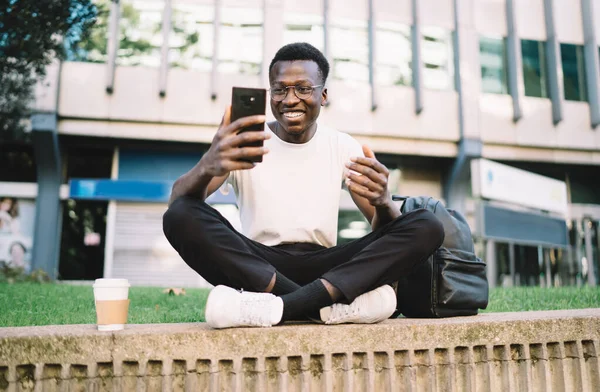 Happy dark skinned hipster guy in casual wear and eyeglasses having fun browsing and chatting online via mobile phone on street
