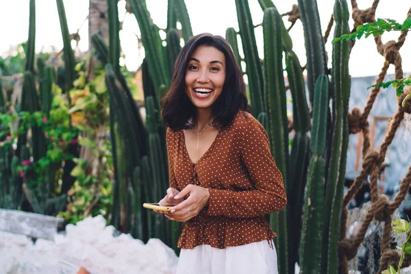 Positive young ethnic female with mobile phone in hands laughing and looking at camera while standing near long cacti in garden