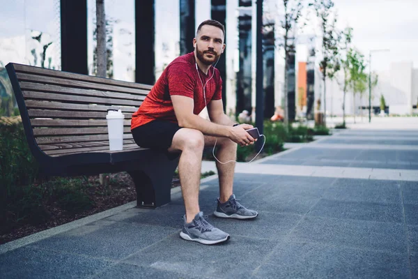 Full body of confident male athlete in earphones sitting on bench and looking away while chilling after morning training in city