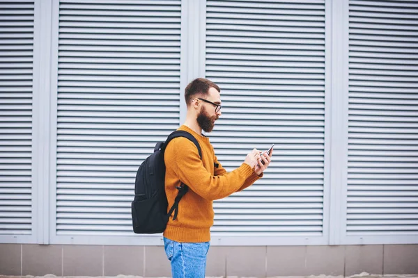 Side view of young bearded hipster with eyeglasses and backpack dialing number on mobile phone beside building with shutter roller curtains on windows
