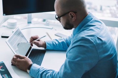 Mavi gömlek ve gözlük takan tanınmamış sakallı işadamının çalışma masasında çalışırken tablete dijital kalemle not alışının yan görüntüsü.