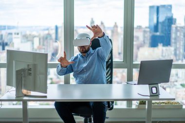 Büyük panoramik pencereli modern ofiste iş sırasında monitör ve dizüstü bilgisayarla masada oturan VR gözlükleri takan iş adamı.