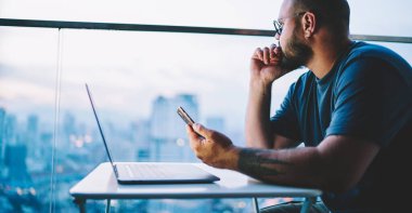 Gündelik giysiler içindeki ciddi bir erkeğin modern iş yerinde çalışırken dizüstü bilgisayarlı ve akıllı telefonlu bir masada oturmasının yan görüntüsü.