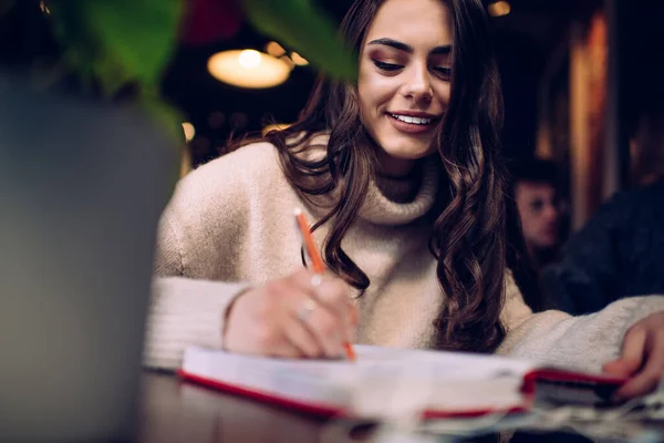 Cheerful young woman with dark hair in casual wear taking notes on notebook and smiling while writing in diary in modern library