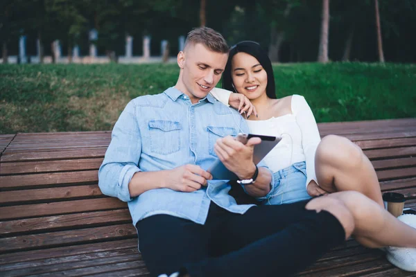 Cheerful Ethnic Couple Casual Wear Sitting Wooden Bench Browsing Tablet — стоковое фото