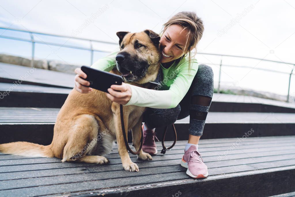 Joyful fit woman having fun with adorable mongrel dog with dark muzzle using cellphone front camera for clicking selfie images