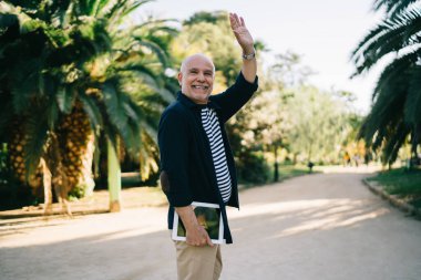 Parkta eğlenirken el sallayan ve gülümseyen modern dijital aletli neşeli erkek turist. 