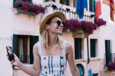 Yazlık elbisesiyle tatmin olmuş bir kadın gülümseyip gözlerini kaçırırken dinlenme yerinde çiçeklerle birlikte siyah akıllı telefon ekranını gösteriyor.