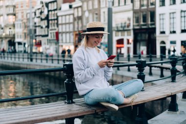 Günlük kıyafetleri ve şapkasıyla neşeli genç bir bayan Amsterdam 'da tatilde dinlenirken ahşap bankta oturuyor ve akıllı telefonla mesajlaşıyor.