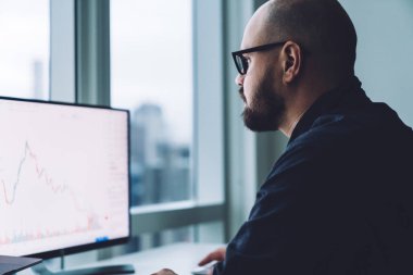 Modern ofiste oturan ve kripto para birimi bitcoin grafikleriyle çalışan resmi giyimli ve gözlüklü bir erkek girişimcinin yan görünüşü