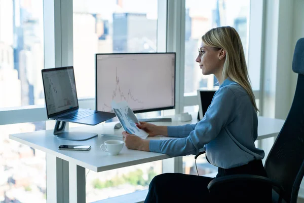 侧视图内容年轻的女性数据分析人员在现代照明办公室工作时 坐在办公桌前 手持现代笔记本电脑和计算机显示器 — 图库照片