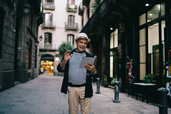 Elderly Caucasian male tourist in electronic headphones making international video call in roaming using modern touch pad and 4g internet