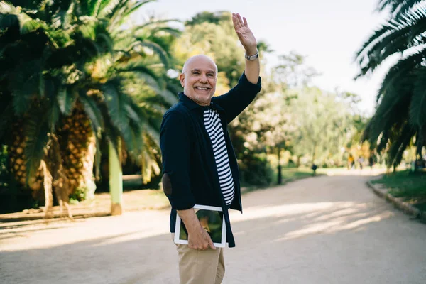 Parkta eğlenirken el sallayan ve gülümseyen modern dijital aletli neşeli erkek turist. 