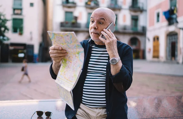 Wonder Caucasian tourist reading travel map searching showplace location and feeling shocked during international cellphone conversation for discussing vacations trip