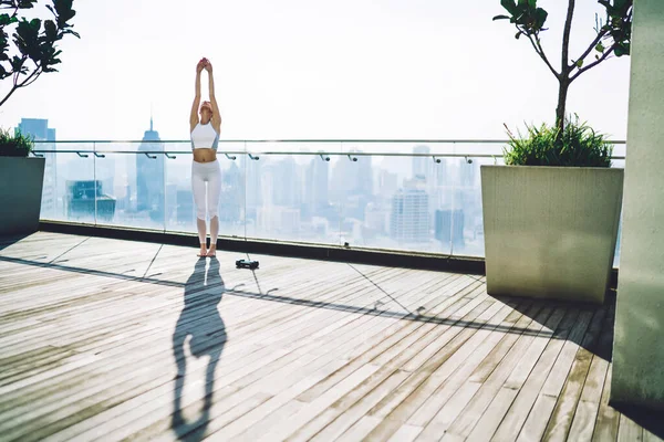 Spor elbiseli bir kadın terasta antrenmandan önce ellerini kaldırarak esneme egzersizi yapıyor ve ısınıyor.