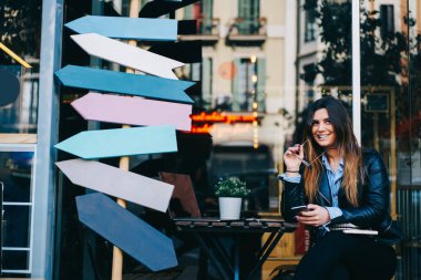 Gülümseyen serbest çalışan bir kadın sokakta tabelaların yanında oturmuş akıllı telefon ve gözlük çiğniyor ve teklif etmeyi düşünüyor.
