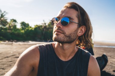 Gözlüklü ve atletli genç bir erkek gezgin, tropik tatil beldesinde tatilin tadını çıkarırken gözlerini kaçırıp sahilde dinleniyor.