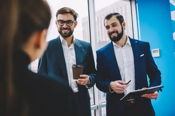 Positive young lawyers in formal suits joking with colleague looking at each other having coffee break in modern office hallway