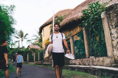 İçeriğin düşük açısı spor giyimli sakallı bir erkek sporcu asfalt yolda sörf tahtası ve spor ayakkabılarıyla geziniyor ve tırmanma tesislerinin yanından geçen evlerin önünden geçiyor.