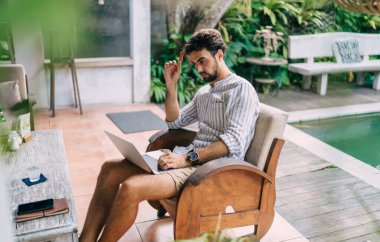 Düşünceli uzaktan kumandalı erkek çalışanın internette internette dolaşırken iş sırasında parmaklarını kaldırarak sandalyeye oturması.