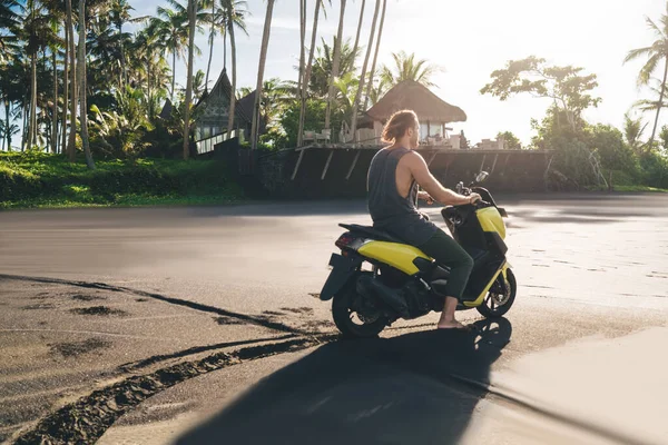 Back view of full body anonymous male driving motorbike and looking away while resting alone in tropical resort and enjoying summer vacation