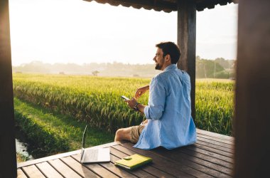 Güneşli bir günde kırsal kesimde dizüstü bilgisayar kullanıp gözlerini kaçıran, günlük kıyafetleriyle gezen mutlu, serbest çalışan bir erkek.