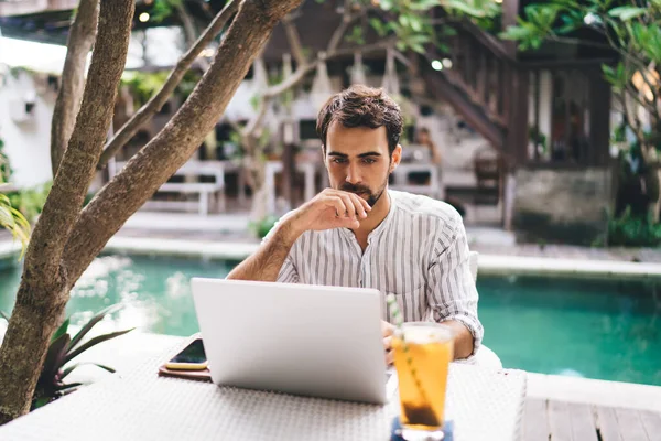 Genç düşünceli sakallı erkek internette gezinirken yazın havuz kenarında terasta bir bardak içkiyle masada kafa patlatıyor.