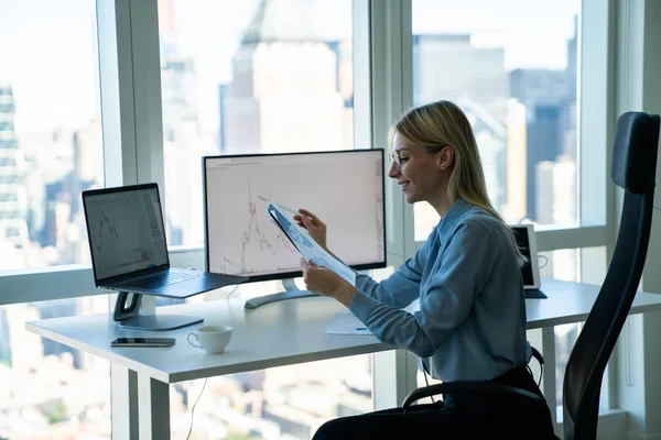 在阅读报告和工作时 身穿正式服装 头戴笔记本电脑和计算机显示器的年轻女企业家坐在办公桌前的侧视图 — 图库照片
