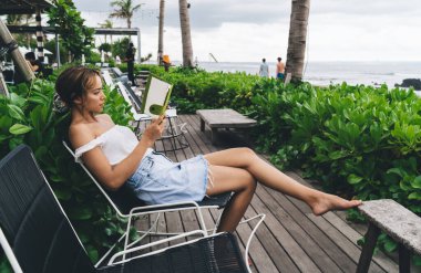 Ciddi etnik kız öğrencilerin yüksek açılı gündelik kıyafetleriyle okyanus kıyısındaki terasta oturuyor ve yaz tatillerinde notlar okuyor.