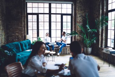 Genç Asyalı kadın pencere kenarında oturuyor ve çalışırken çeşitli meslektaşlarıyla yaratıcı çatı katında bilgisayar kullanarak gülüyor.