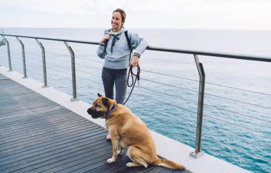 Rıhtımda duran, korkuluklara yaslanan genç, pozitif bir kadın. Deniz kenarında yürürken köpek tasmanın yanında oturuyor.