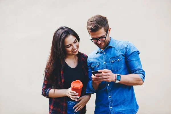 Cheerful female student in casual outfit with takeaway cup standing near joyed male friend in glasses showing information on smartphone against beige background