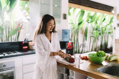 Masanın yanında durup sağlıklı sebzeli salata hazırlayan kahverengi saçlı, şık beyaz elbiseli çekici Asyalı kadın.