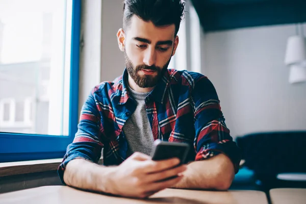 Ciddi genç hipster adam kampüste otururken akıllı telefonuyla para transferi yapıyor. Milenyum erkeği sosyal ağlarda cep telefonu haberlerini içeride kablosuz internet üzerinden kullanıyor..