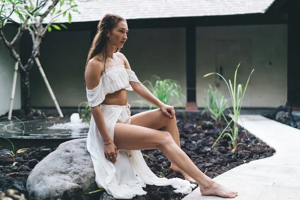 Beyaz giyimli, çıplak ayaklı, stil sahibi bir kadın taşın üzerinde oturuyor ve bahçede dinlenirken yüzünü çeviriyor.