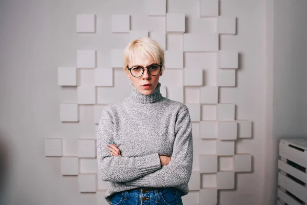Serious woman in casual gray sweater and jeans with folded hands looking at camera spending time in modern light room