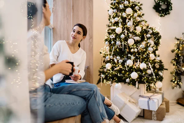 Young female friends in casual outfits sitting on windows sill near decorated Christmas tree and drinking cup of coffee in living room