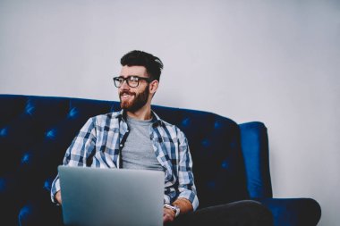 Modern dizüstü bilgisayar ile rahat kanepe oturma ve e öğrenme, başarılı hipster adam Eyewear bir kenara netbook üzerinde çalışmanın tatilinde yüzünde sevimli gülümseme ile seyir zevk mutlu beyaz blogger